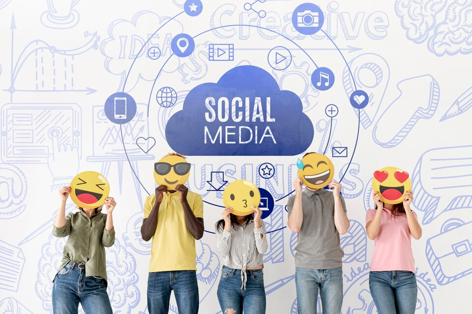 five people holding emoji face cutouts stand in front of a wall with social media icons and text